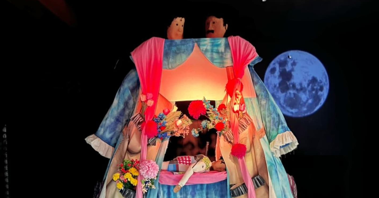 Títeres forman un teatrillo que es un vestido o traje y en su interior, en el escenario del teatrillo, hay otro títere más pequeño tumbado. 